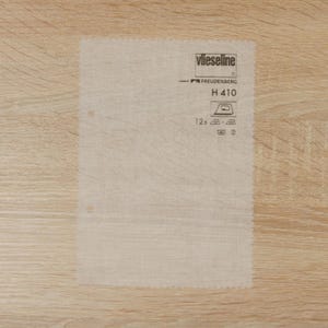 Peut inclure: Une feuille rectangulaire de tissu thermocollant Vlieseline H 410. La feuille est de couleur beige clair et présente les logos Vlieseline et Freudenberg, ainsi que des symboles d'entretien pour le repassage et le lavage. Deux petits trous sont visibles.