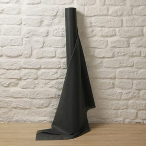 Puede incluir: Un rollo de tela gris oscuro, parcialmente desenrollado, muestra su textura. Se encuentra contra una pared de ladrillos blancos y descansa sobre una superficie de madera. La estética es simple y funcional, ideal para proyectos de manualidades.