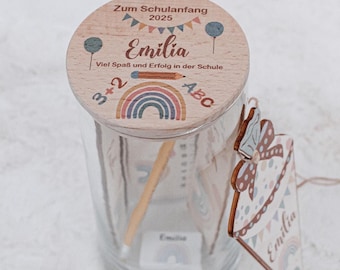 May include: A wooden jar with a lid decorated with a rainbow, balloons, and the text "Zum Schulanfang 2025 Emilia Viel Spaß und Erfolg in der Schule 3+2 ABC". The jar is filled with pencils and a small wooden tag with the text "Emilia" is attached to the jar.
