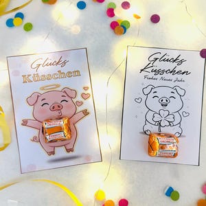 Könnte beinhalten: Zwei Grußkarten mit Schweine-Illustrationen und deutschem Text, "Glücks Küsschen" und "Frohes Neues Jahr". Jede Karte hält eine verpackte Schokolade. Die Szene ist mit Konfetti und goldenen Bändern dekoriert.