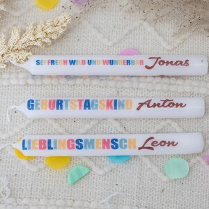 Peut inclure: Trois bougies d'anniversaire blanches avec du texte coloré et des noms. La bougie du haut indique "Sei frech wild und wunderbar Jonas". La bougie du milieu indique "Geburtstagskind Anton". La bougie du bas indique "Lieblingsmensch Leon".
