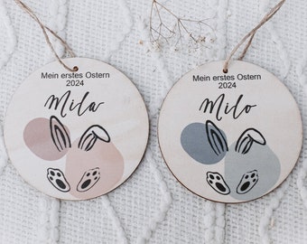 Peut inclure: Deux décorations de Pâques en bois avec des oreilles de lapin et des empreintes de pas. L'une est rose et l'autre est bleue. Le texte sur les ornements indique "Mein erstes Ostern 2024" et les noms "Mila" et "Milo".