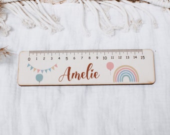 May include: A wooden ruler featuring a rainbow, balloon, and bunting design. The ruler is marked with numbers from 0 to 15. The name "Amelie" is written in cursive on the ruler.
