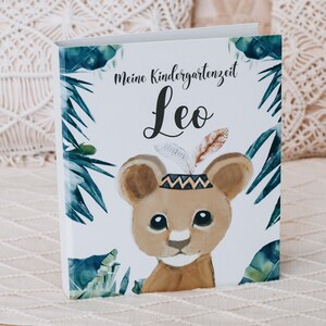 Puede incluir: Un libro blanco con un tema de jungla que presenta un lindo cachorro de león con un tocado. El título del libro es "Meine Kindergartenzeit Leo".