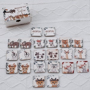 Könnte beinhalten: Ein Holz-Memory-Spiel mit Dschungel-Motiv. Das Spiel enthält 16 Paare von Spielsteinen mit verschiedenen Tieren, darunter Faultiere, Pinguine, Pandas, Hirsche, Koalas und Löwen. Die Spielsteine sind mit floralen Akzenten verziert und der Text "Sofia" erscheint auf zwei der Spielsteine.