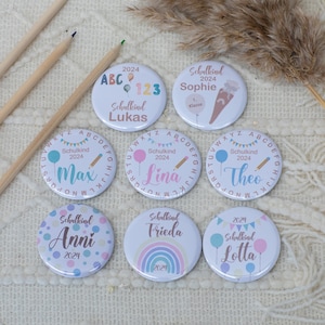 Op de afbeelding: Negen kleurrijke ronde knoppen met verschillende ontwerpen en tekst. De knoppen hebben verschillende thema's, waaronder ballonnen, potloden en alfabetletters. De tekst op de knoppen bevat namen zoals "Lukas", "Sophie", "Max", "Lina", "Theo", "Anni", "Frieda" en "Lotta". De knoppen bevatten ook het jaar "2024" en het woord "Schulkind", wat Duits is voor "schoolkind".