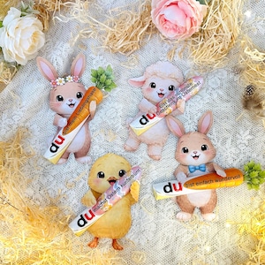 Peut inclure: Quatre découpes en papier sur le thème de Pâques représentant un lapin, un agneau et un poussin tenant des carottes avec du texte allemand. Le lapin porte une couronne florale et l'agneau a un pelage blanc duveteux.