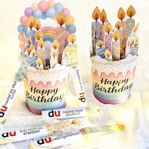 Może przedstawiać: Dekoracje tortu urodzinowego w stylu akwarelowym ze świecami i napisem "Happy Birthday". Ciasta są ozdobione pastelowymi kolorami i tęczowymi akcentami. Obecnych jest również kilka małych kart w kształcie świec z niemieckim tekstem.