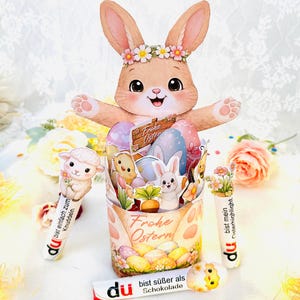 May include: An Easter-themed display with a large, smiling bunny wearing a floral crown. The pop-up scene features Easter eggs, bunnies, and a chick. Includes decorative pens and a chocolate bar with text in German.