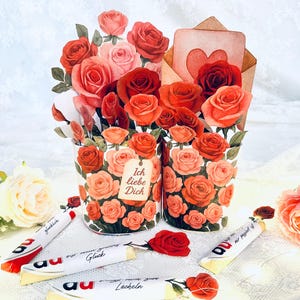 Puede incluir: Un arreglo floral romántico con rosas de papel en tonos rojos, rosas y naranjas. El arreglo incluye dos jarrones decorativos, una tarjeta con un corazón y varios caramelos envueltos. El texto "Ich liebe Dich" es visible.