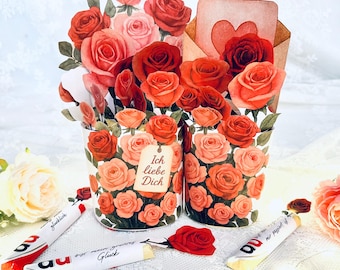 Pode incluir: Um arranjo floral romântico com rosas de papel em tons de vermelho, rosa e laranja. O arranjo inclui dois vasos decorativos, um cartão com um coração e vários doces embalados. O texto "Ich liebe Dich" é visível.