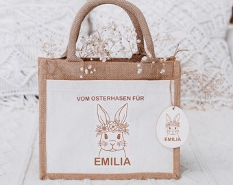 Może przedstawiać: Brązowa torba jutowa z białą podszewką z tkaniny. Torba ma białą metkę z ilustracją królika i imieniem "EMILIA". Torba ma również biały panel z tkaniny z ilustracją królika i tekstem "VOM OSTERHASEN FÜR EMILIA".