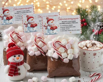 May include: A Christmas-themed image showing bags of hot chocolate mix with marshmallows and candy canes. A snowman toy and a mug of hot chocolate with marshmallows are also visible. The bags have recipe cards that read "Schneemannsuppe".