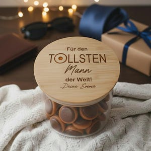 Könnte beinhalten: Ein Glas mit Schokoladenbonbons, bedeckt mit einem Holzdeckel. Der Deckel ist mit deutschem Text graviert. Weitere Gegenstände im Hintergrund sind eine Brieftasche, eine Sonnenbrille, ein Geschenk mit blauem Band und eine blaue Krawatte.