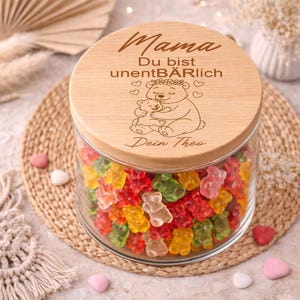 Pode incluir: Um frasco de vidro cheio de gomas coloridas em forma de urso. O frasco tem uma tampa de madeira com texto gravado e uma ilustração de urso. O texto na tampa diz "Mama, Du bist unentBÄRLich, Dein Theo."