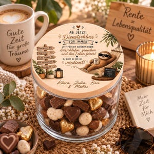 Könnte beinhalten: Ein klares Glasgefäß gefüllt mit gemischten Pralinen, mit einem runden Holzdeckel, der eine Illustration eines Faultiers in einer Hängematte, deutschen Text sowie die Namen Klara, Julia, Ben und Matteo zeigt, umgeben von Dekorationsartikeln.