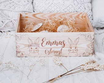 Może przedstawiać: Drewniany kosz wielkanocny z napisem "Emmas Osternest" oraz ilustracjami królików i kwiatowych wzorów. Kosz wypełniony jest słomą i drewnianymi dekoracjami wielkanocnymi.