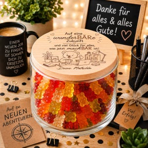 May include: A clear glass jar filled with colorful gummy bears, topped with a round wooden lid engraved with a bear illustration, the text 'Auf eine wunderBÄRe Zukunft', and the name 'Deine Mathilda' alongside office-themed graphics.