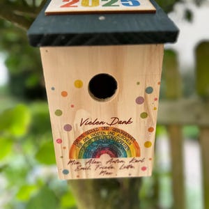 May include: A wooden birdhouse with a dark green roof and the year "2025" in colorful letters. The birdhouse has a round entry hole and is decorated with colorful polka dots and text in German.