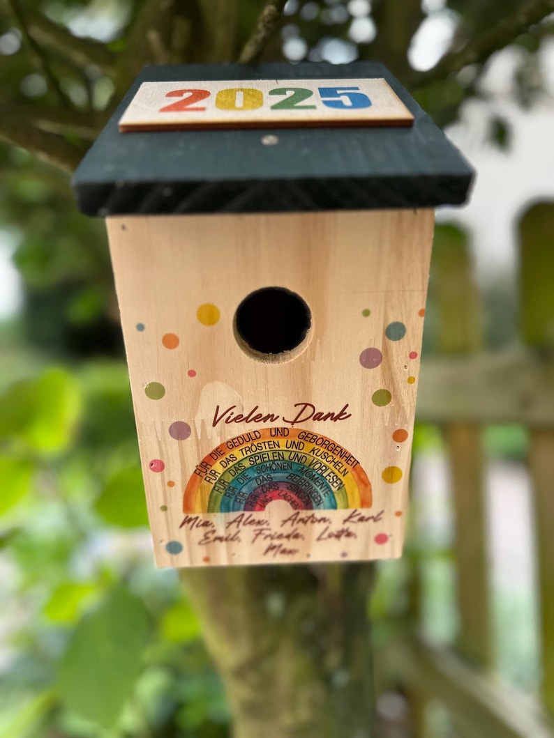 May include: A wooden birdhouse with a dark green roof and the year "2025" in colorful letters. The birdhouse has a round entry hole and is decorated with colorful polka dots and text in German.
