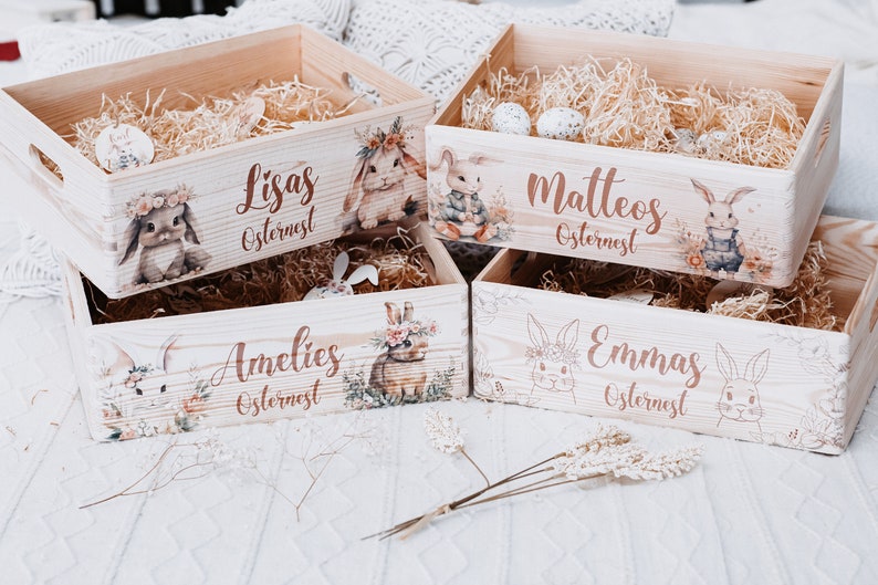 Puede incluir: Cuatro cajas de Pascua de madera con ilustraciones de conejos y nombres: Lisas, Matteos, Amelies y Emmas. Cada caja est&aacute; llena de paja y decorada con detalles florales. Las cajas son de color marr&oacute;n claro.
