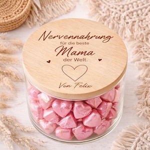 Pode incluir: Um frasco de vidro cheio de doces rosa em forma de coração, coberto com uma tampa de madeira. A tampa é gravada com as palavras "Nervennahrung für die beste Mama der Welt" e um símbolo de coração, com o nome "Von Felix" abaixo.