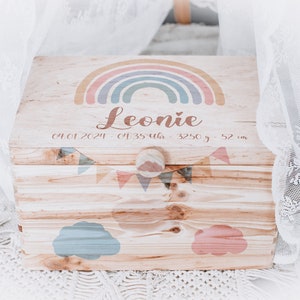 Könnte beinhalten: Eine Holzkiste mit einem Regenbogen-, Wolken- und Girlanden-Design. Die Kiste ist mit dem Namen "Leonie" und dem Datum "04.01.2021" sowie der Uhrzeit, dem Gewicht und der Größe des Babys personalisiert.