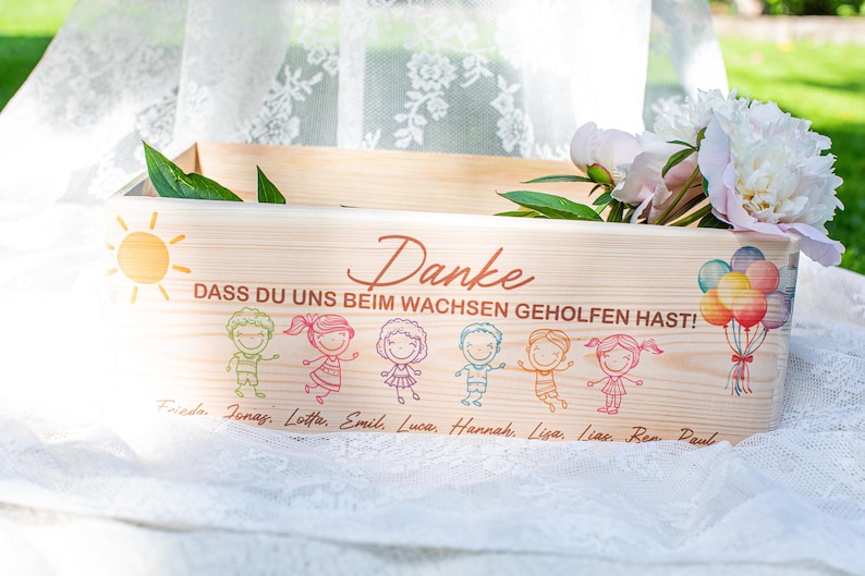 May include: A wooden box with the German word "Danke" and the phrase "DASS DU UNS BEIM WACHSEN GEHOLFEN HAST!" printed on it. The box is decorated with cartoon children, a sun, and balloons. Peonies and greenery are inside the box.