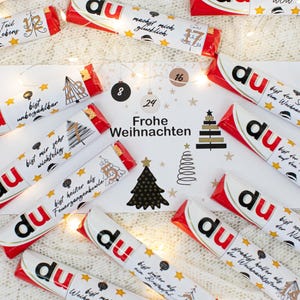 May include: An advent calendar featuring chocolate bars. The bars are white with red detailing and the word "du" in black and red. The bars are decorated with text and illustrations. A central card displays "Frohe Weihnachten" and Christmas tree designs.