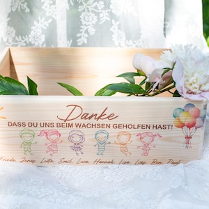 May include: A wooden box with a painted sun, balloons, and children's figures. The box has the word "Danke" and the phrase "DASS DU UNS BEIM WACHSEN GEHOLFEN HAST!" printed on it. A bouquet of flowers rests on top.