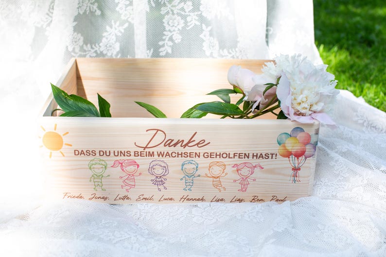 May include: A wooden box with a painted sun, balloons, and children's figures. The box has the word "Danke" and the phrase "DASS DU UNS BEIM WACHSEN GEHOLFEN HAST!" printed on it. A bouquet of flowers rests on top.