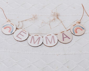 Peut inclure: Une bannière en bois avec le nom "EMMA" écrit en lettres roses. Chaque lettre est sur une pièce de bois ronde séparée. La bannière est enfilée avec de la ficelle et présente deux motifs d'arc-en-ciel aquarelle aux extrémités.