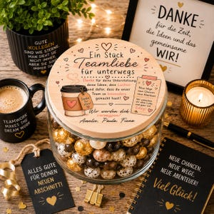 Op de afbeelding: Een doorzichtige glazen pot gevuld met diverse chocoladeballetjes, afgesloten met een rond houten deksel met een illustratie van een koffiebeker, hartjes en Duitse tekst waaronder "Ein Stück Teamliebe für unterwegs" en de namen Amelie, Paula, Fiona.