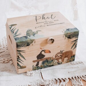 Può includere: Una scatola di legno con un tema giungla. La scatola è decorata con un tucano, un bradipo e foglie tropicali. Il testo "Phil, Meine kleine Box mit den tollen Erinnerungen" è stampato sul coperchio.