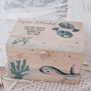 Può includere: Una scatola portaoggetti in legno con una finitura in legno chiaro e un design a tema oceanico blu e bianco. La scatola ha un coperchio incernierato e una piccola manopola sul davanti. Il coperchio è decorato con una balena, due pesci, una stella marina e una tartaruga marina. La scatola ha anche il testo "Kaden Kittelchok" e "16.03.2018 06:41 Uhr 3350 g 51 cm" sul coperchio.