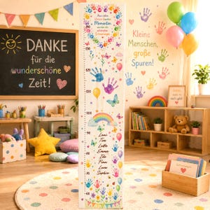 Pode incluir: Uma régua de crescimento vertical em madeira com uma escala de 60 a 140 cm, decorada com impressões de mãos coloridas, borboletas, balões, um arco-íris e o texto em alemão: Aus vielen kleinen bunten Momenten wurden die schönsten Erinnerungen.