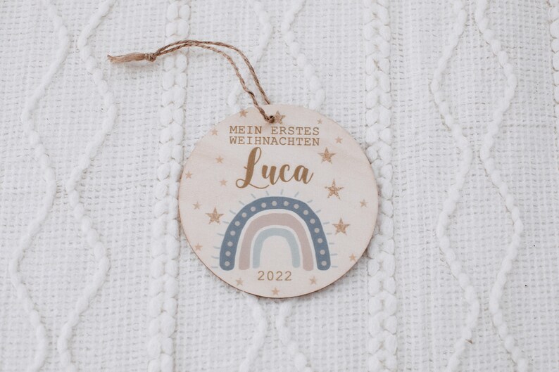 K&ouml;nnte beinhalten: Ein runder Holzanh&auml;nger mit einem Regenbogen, Sternen und dem Text "Mein erstes Weihnachten Luca 2022".