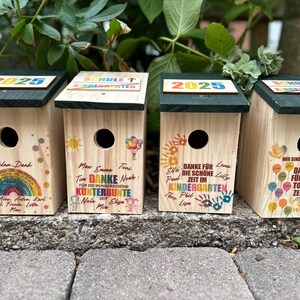 May include: Four wooden birdhouses with dark green roofs, each featuring unique colorful designs and text. One has a rainbow, another has balloons, and two have handprints. The text includes "2025" and "Kindergarten."