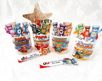 May include: A collection of colorful bear-shaped superhero figurines in white cups with German text. The bears wear capes and masks. The cups have phrases like "Papa, you are my superhero" and "Thank you, Dad." Also included are white and red candy bars.
