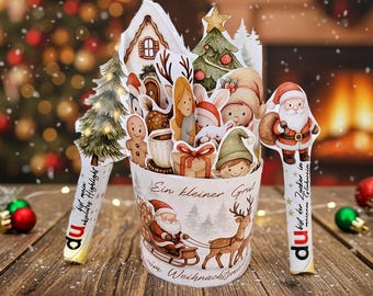 May include: A festive Christmas-themed papercraft display. The centerpiece is a decorated bucket with Santa Claus and reindeer imagery. It features various cut-out figures, including a gingerbread man, elf, and gift boxes. The text "Ein kleiner Gruß" is visible.