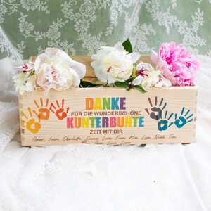 May include: A wooden box with colorful handprints and the German text "DANKE F&Uuml;R DIE WUNDERSCH&Ouml;NE KUNTERBUNTE ZEIT MIT DIR." The box is filled with white and pink peonies. The box is on a white lace surface.