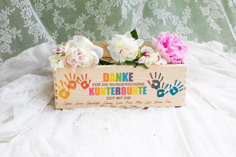 May include: A wooden box with colorful handprints and the German text "DANKE F&Uuml;R DIE WUNDERSCH&Ouml;NE KUNTERBUNTE ZEIT MIT DIR." The box is filled with white and pink peonies. The box is on a white lace surface.