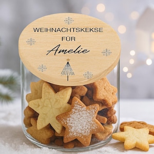 Può includere: Un barattolo di vetro pieno di biscotti a forma di stella, alcuni spolverati di zucchero a velo. Il coperchio di legno è inciso con la scritta "WEIHNACHTSKEKSE FÜR Amelie" e decorato con fiocchi di neve e un albero di Natale.