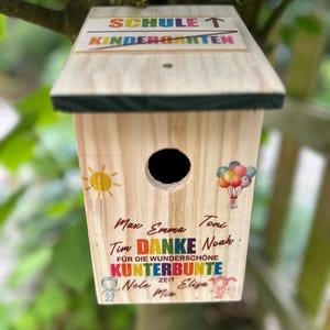 May include: A wooden birdhouse with a sloped roof, featuring the text "SCHULE T KINDERGARTEN". The birdhouse has a round entry hole and is decorated with a sun, balloons, and colorful text including names and the word "DANKE".