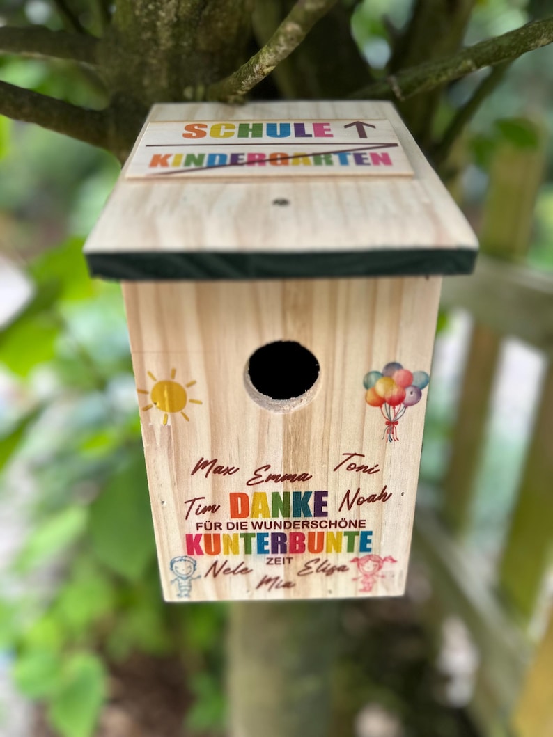 May include: A wooden birdhouse with a sloped roof, featuring the text "SCHULE T KINDERGARTEN". The birdhouse has a round entry hole and is decorated with a sun, balloons, and colorful text including names and the word "DANKE".