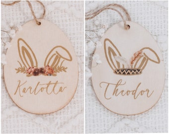 Op de afbeelding: Twee houten paaseieren met konijnenoren. Een ornament is versierd met bloemen en heeft de tekst "Karlotta". De andere ornament heeft een tribale ontwerp en heeft de tekst "Theodor".