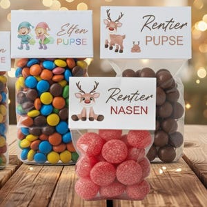 May include: Clear cellophane bags filled with colourful sweets and chocolates, each topped with a white label featuring festive illustrations and text. The labels read "Elfen PUPSE" and "Rentier PUPSE" and "Rentier NASEN". The sweets are arranged on a wooden surface.