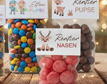 Puede incluir: Bolsas transparentes de celofán llenas de caramelos y chocolates de colores, cada una coronada con una etiqueta blanca con ilustraciones y texto festivos. Las etiquetas dicen "Elfen PUPSE", "Rentier PUPSE" y "Rentier NASEN". Los caramelos están dispuestos sobre una superficie de madera.