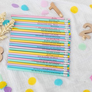 Puede incluir: Un conjunto de 20 l&aacute;pices de colores pastel con mensajes de &aacute;nimo impresos en ellos. Los l&aacute;pices est&aacute;n dispuestos en una pila y est&aacute;n rodeados de confeti de colores.