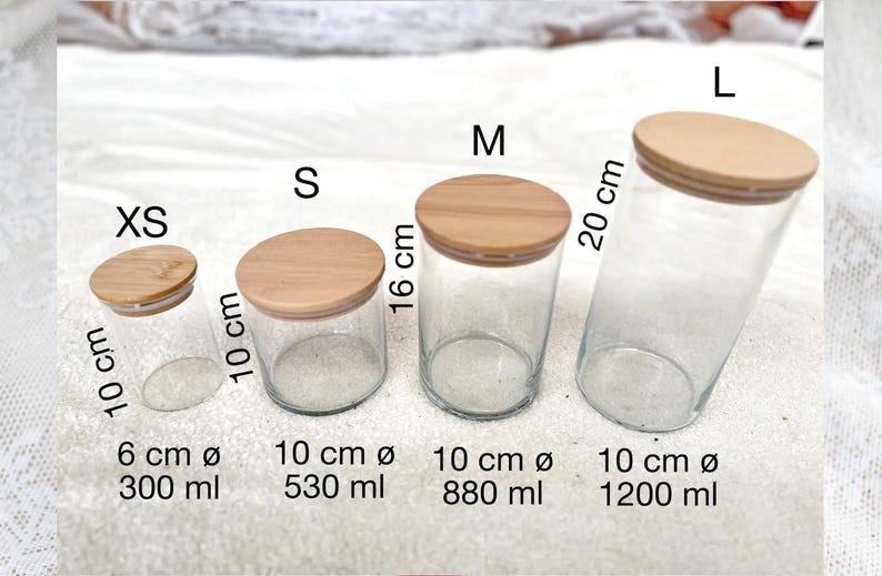 May include: A set of four clear glass jars with wooden lids, labeled XS, S, M, and L. The XS jar is 10 cm tall and 6 cm in diameter, holding 300 ml. The S jar is 10 cm tall and 10 cm in diameter, holding 530 ml.